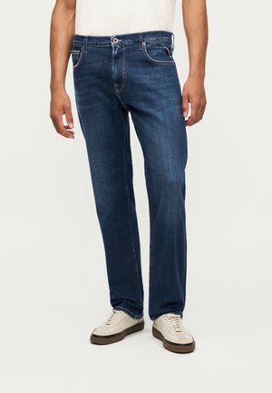 Homme portant un jean droit bleu foncé et des baskets décontractées écrues, debout devant un fond uni clair.
