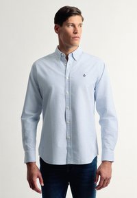 Camisa de manga larga a rayas azul claro y blanca con un cuello clásico y un pequeño logo negro en el pecho. La tela parece suave.