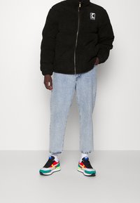 Persona che indossa una giacca in pile nera, jeans azzurri corti, calzini bianchi e scarpe da ginnastica multicolori, in piedi davanti a uno sfondo semplice.