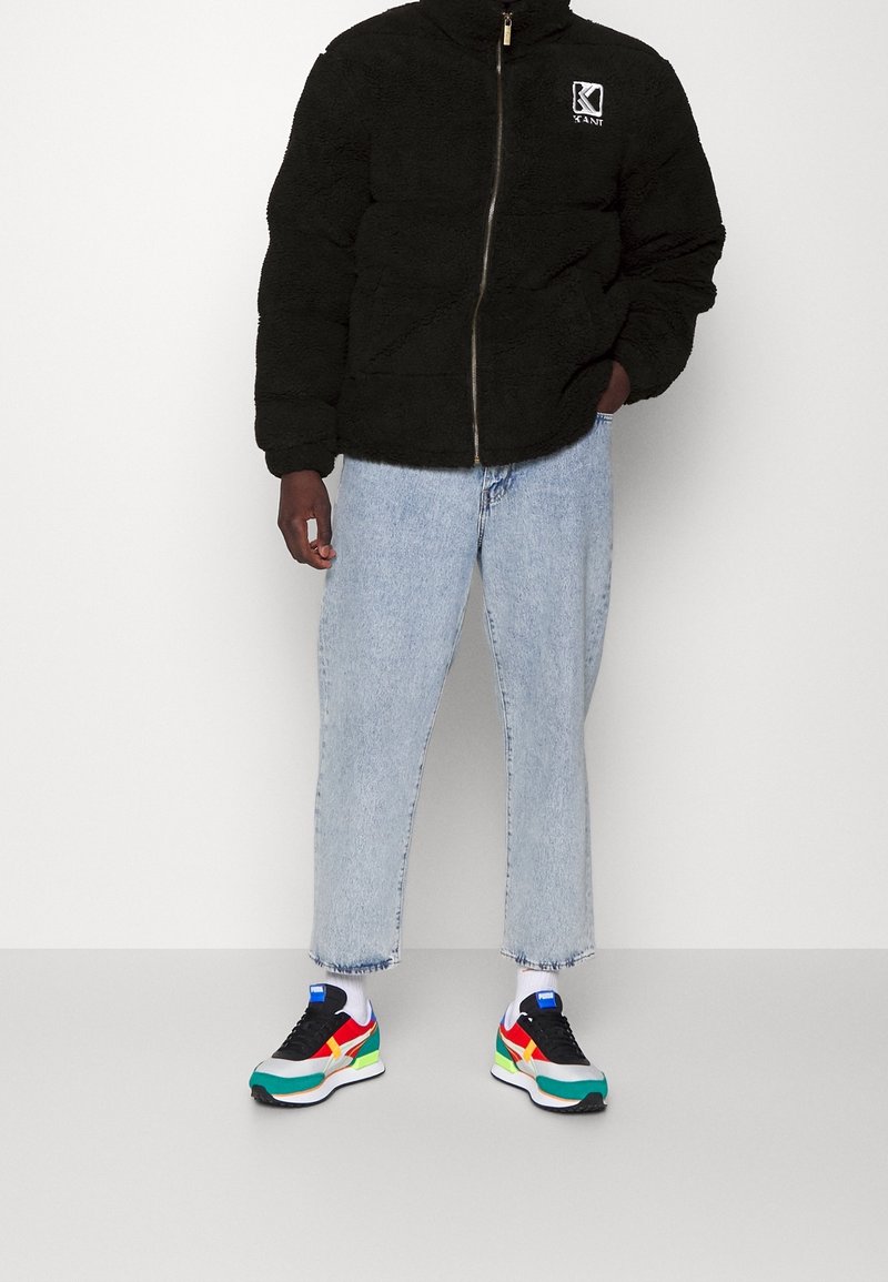 Persona che indossa una giacca in pile nera, jeans azzurri corti, calzini bianchi e scarpe da ginnastica multicolori, in piedi davanti a uno sfondo semplice.