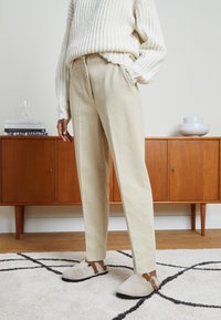Beige tapse broek gecombineerd met een chunky witte geribbelde trui en lichtgrijze instappers met bruine accenten, op een zacht gemêleerd tapijt.