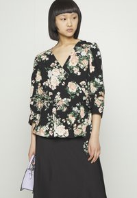 Blusa florinda preta com decote em V e mangas três-quartos. Apresenta rosas pêssego e folhas verdes numa textura de tecido. Combinada com uma saia preta.