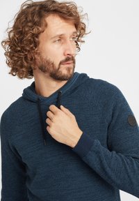 Dunkelblauer, strukturierter Kapuzenpullover mit einem Schalkragen, schwarzen Kordeln und einem runden Logo-Patch auf der linken Schulter. Gerippte Bündchen und Saum.