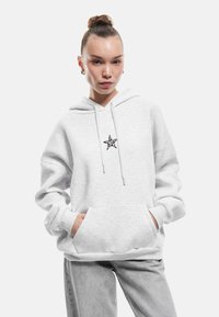 Lichtgrijze hoodie van een zachte katoenmix met een voorzak. Bevat een zwart sterrenlogo op de borst en verstelbare trekkoorden.