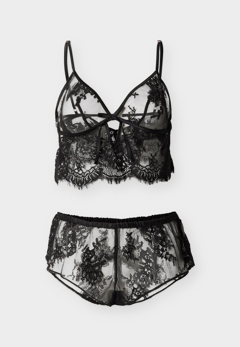 Μαύρο δαντελένιο σουτιέν bralette με ρυθμιζόμενες λεπτές τιράντες και δέσιμο μπροστά, συνδυασμένο με αντίστοιχα σορτς από δαντέλα ψηλόμεσα, και τα δύο διάφανα με λουλουδάτα σχέδια.