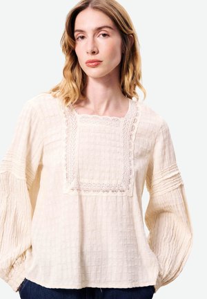 Femme aux cheveux blonds mi-longs portant un chemisier crème texturé avec des détails en dentelle et des manches longues bouffantes sur un fond uni.