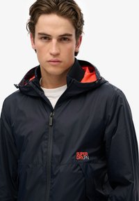 Chaqueta ligera de marina con cierre de cremallera, capucha y acentos en naranja; presenta un logo en el lado izquierdo del pecho y una textura suave, resistente al agua.