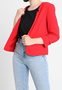 Blazer rouge en tissu texturé, avec une coupe ajustée, un devant ouvert et des manches trois-quarts, associé à un haut noir et un jean bleu.