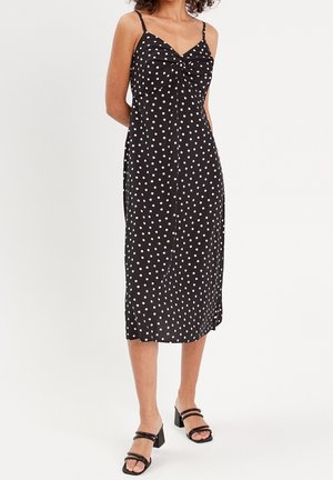 Femme portant une robe midi noire à pois blancs, avec des bretelles spaghetti, un détail torsadé à l'avant et des sandales à talons noires.