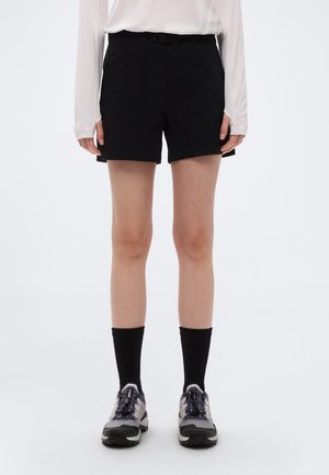 Person iført sorte shorts, sorte crew-sokker, grå og sorte sneakers og en hvid bluse med lange ærmer, stående foran en ensfarvet baggrund.