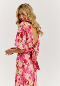 Šaty bez zadního dílu s květinovým vzorem, které mají růžový základ s červenými, zelenými a krémovými motivy, krátké rukávy a zavazovací ozdobu v pase.