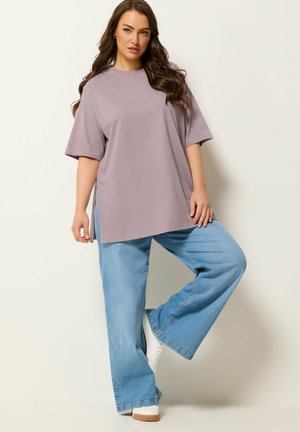 Vrouw met lang golvend haar, gekleed in een losvallend mauve T-shirt, lichtblauwe wijde jeans en witte schoenen, staand met één been omhoog.