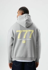 Grijze hoodie met een groot geel nummer "777" en tekst "WORLD IS YOURS" op de achterkant. Voorzien van een capuchon met trekkoorden en geribbelde zoom.