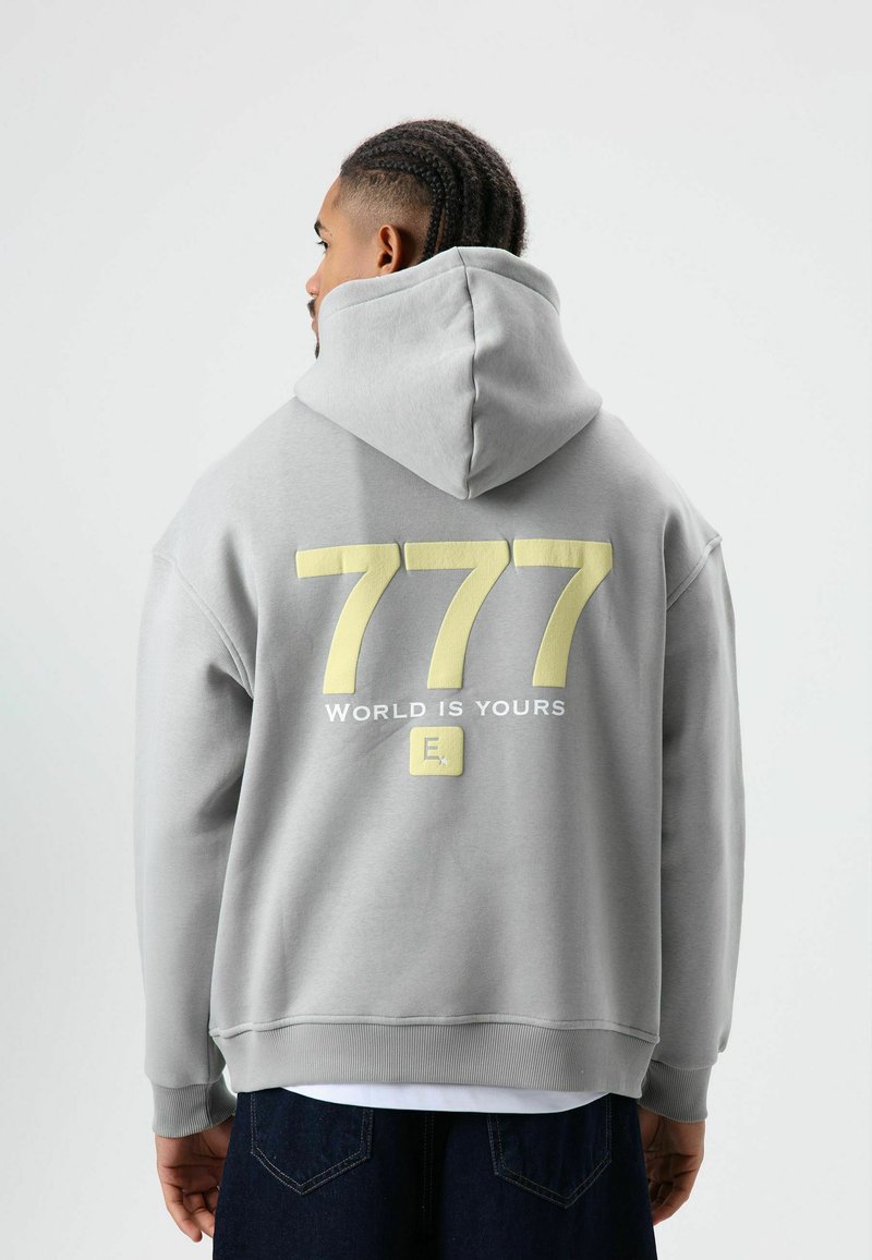 Grijze hoodie met een groot geel nummer "777" en tekst "WORLD IS YOURS" op de achterkant. Voorzien van een capuchon met trekkoorden en geribbelde zoom.