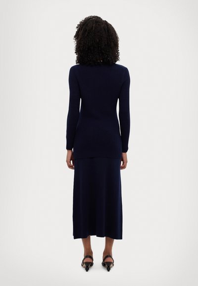 Maglione a coste blu navy con maniche lunghe, abbinato a una lunga gonna blu navy che arriva a metà polpaccio, indossato con scarpe col tacco nere.