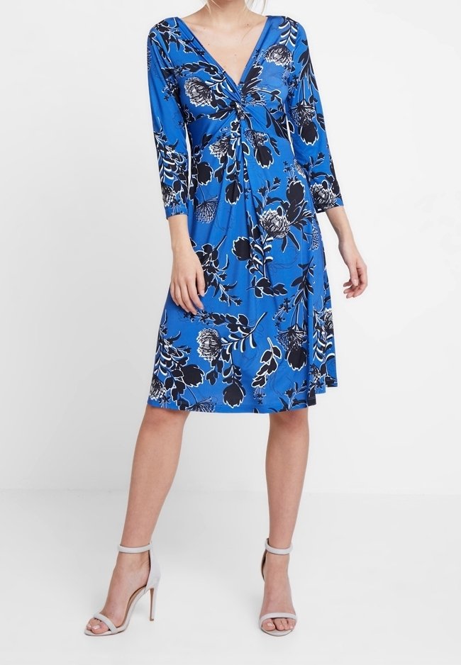 Donna che indossa un vestito blu al ginocchio con un motivo floreale bianco e nero, scollatura a V, maniche a tre quarti e sandali bianchi con tacco alto.