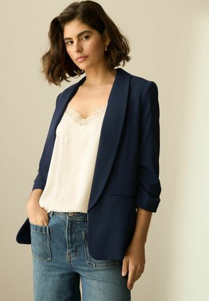Marineblazer med en afslappet pasform, to lommer og opsmøgte ærmer, båret over en cremefarvet top med pailletkant og højtaljede denimjeans.