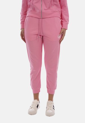 Donna che indossa pantaloni da jogging rosa e felpa con cappuccio abbinata con cerniera, con scarpe da ginnastica bianche, in piedi contro uno sfondo chiaro uniforme.