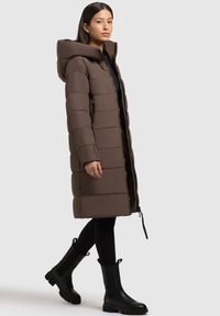 Braune Daunenjacke mit Kapuze, mit gestepptem Design, Reißverschluss, Seitentaschen und knielangem Schnitt. Getragen mit schwarzen Stiefeletten.