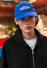 Gorra de béisbol azul con letras blancas que dicen "MÓDULO", llevada con una sudadera negra con cremallera sobre una camiseta blanca. Collar de cadena plateado visible.