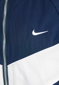 Námořnický a bílý kabát se zipem v přední části. Obsahuje bílou logo Nike, je vyroben z lehkého materiálu s hladkou texturou a designem v blokových barvách.