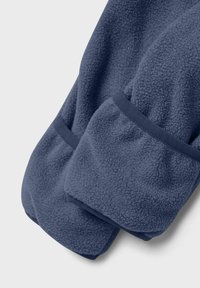 Detalle de la manga de una chaqueta de forro polar azul marino con tejido texturizado y puños cosidos sobre un fondo gris claro.