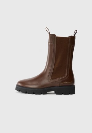 Brązowy skórzany botek za kostkę z elastycznymi panelami bocznymi, czarną podeszwą z bieżnikiem, pętelkami do ściągania i małym logo Tommy Hilfiger w okolicy pięty.