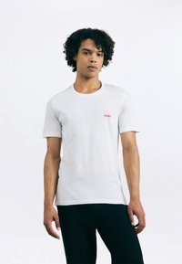 Bărbat tânăr cu părul creț, îmbrăcat cu un tricou alb de la marca Hugo și pantaloni negri, stând în fața unui fundal alb simplu.