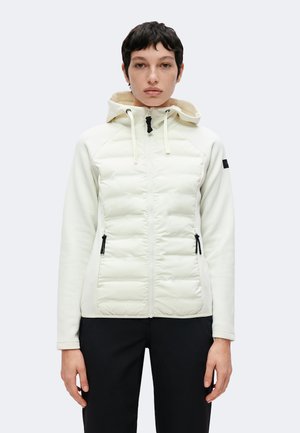 Personne aux cheveux courts et foncés portant une veste matelassée blanche à capuche et un pantalon noir, debout devant un fond clair uni.