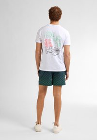 T-shirt bianca in cotone con una grafica colorata di palme e un camion sul retro, abbinata a short da bagno scuri con fantasia.