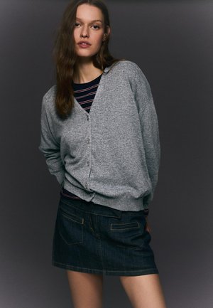 Junge Frau trägt eine graue, geknöpfte Strickjacke über einem gestreiften Hemd und einem dunklen Denim-Minirock, steht vor einem dunklen Hintergrund.