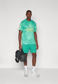 Green mesh sports jersey with "ADIDAS" logo, paired with matching shorts. Black duffel bag held in one hand, white sneakers on feet.