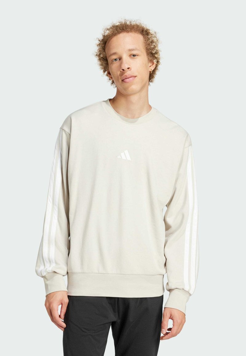 Lys beige sweatshirt lavet af blødt materiale, med hvidt Adidas-logo og tre hvide striber på ærmerne. Lange ærmer, design med rund hals.