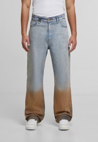 Vyras, vilkintis šviesiai mėlynus džinsus, kurie apatinėje dalyje turi rudos gradientinės spalvos dažymą, baltus sportbačius ir baltą beviršį marškinėlį prieš paprastą foną.