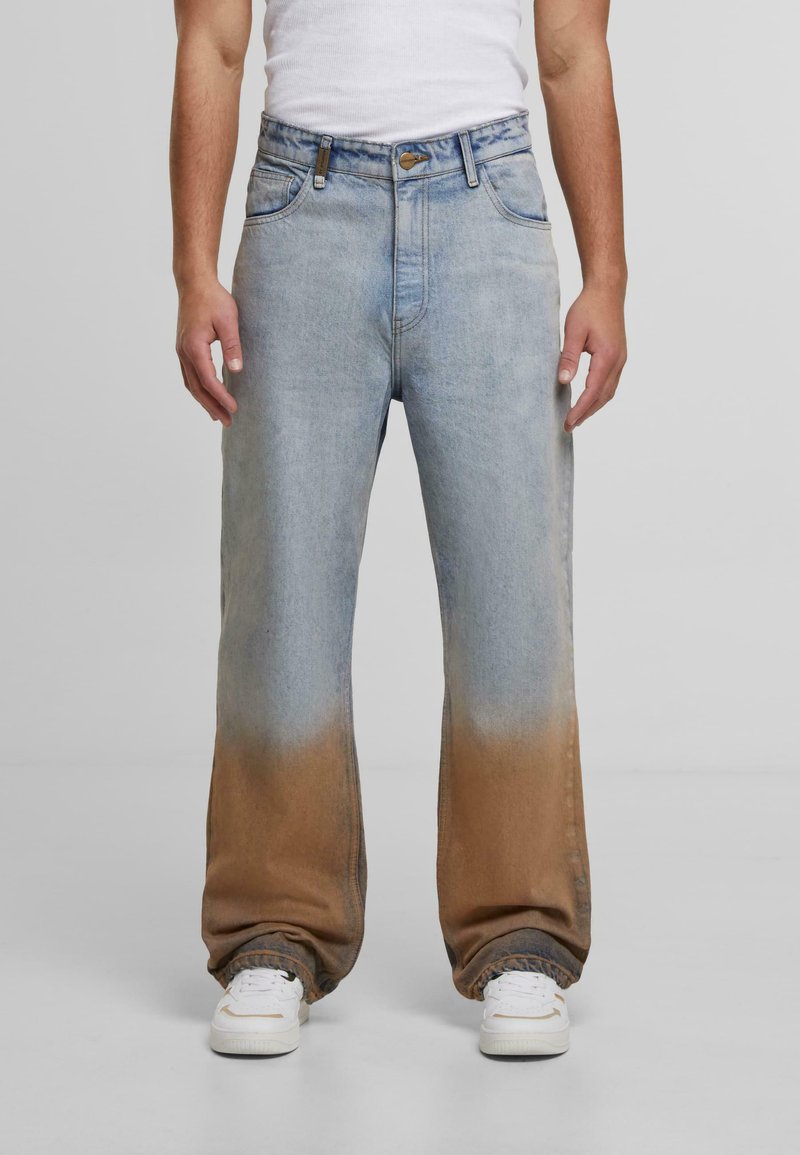 Vyras, vilkintis šviesiai mėlynus džinsus, kurie apatinėje dalyje turi rudos gradientinės spalvos dažymą, baltus sportbačius ir baltą beviršį marškinėlį prieš paprastą foną.