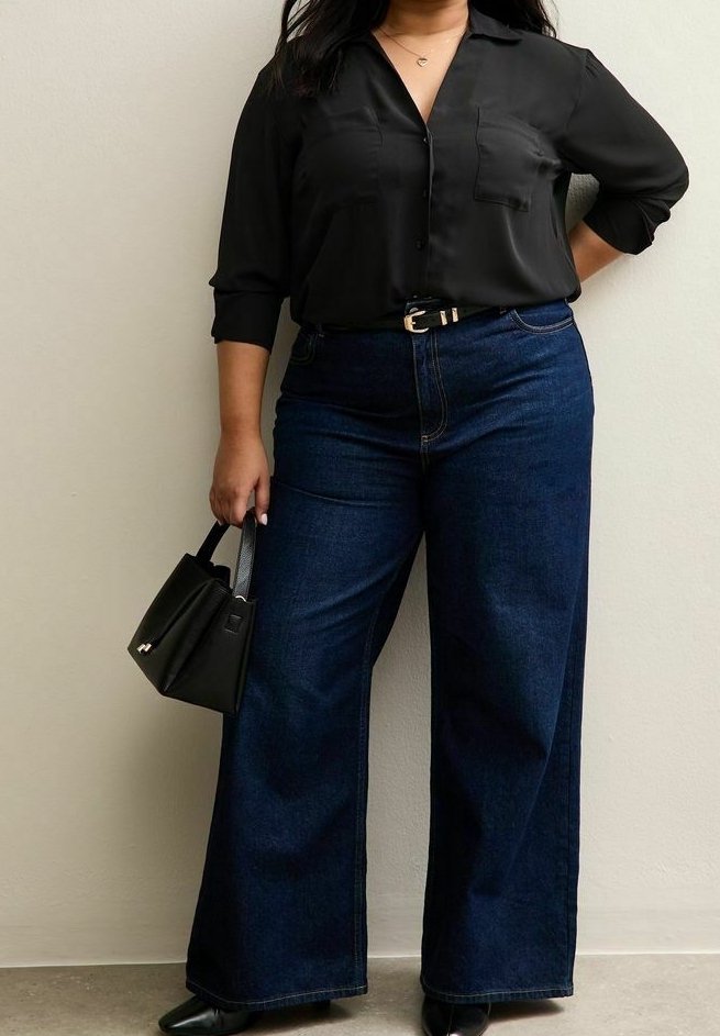 Femme portant une chemise noire boutonnée et un jean large bleu foncé, tenant un petit sac à main noir, debout contre un mur uni.