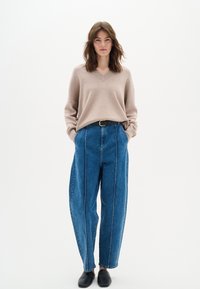 Beżowy sweter w serek, luźny krój. Niebieskie dżinsowe spodnie z wysokim stanem, z białym przeszyciem po bokach, zwężane nogawki. Czarny pasek, czarne buty wsuwane.