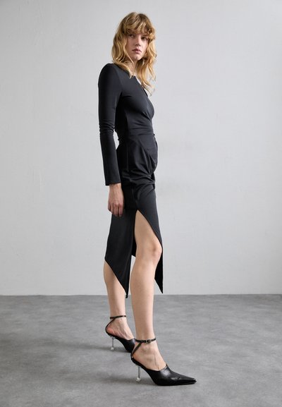 Vestido negro de manga larga con un corsé ajustado y una abertura lateral, combinado con unos zapatos de tacón negro tipo slingback con un acento metálico y punta afilada.
