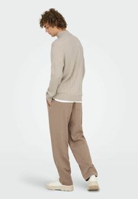 Mann in beigem, weichem Strickpullover, braunen, lockeren Hosen und weißen Sneakers steht mit Händen in den Taschen und blickt nach links unten.