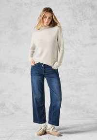 Hellgrauer, gerippter Pullover mit hohem Kragen, kombiniert mit weit geschnittenen, dunkelblauen Jeans. Schuhe: cremfarbene Sneaker mit hellen Sohlen.