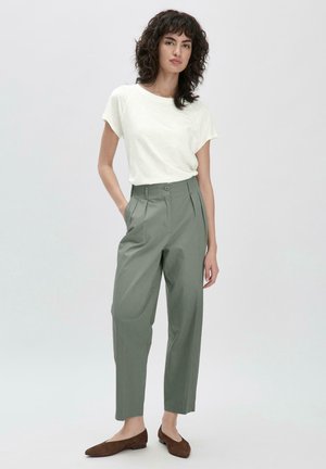 Lichtgrijs T-shirt met korte mouwen; hooggewaisted, geplooide, olijfgroene broek; model staat met één hand in de zak, draagt bruine flats.