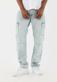Pantalones cargo azul claro de mezclilla con múltiples bolsillos, un ajuste holgado y un ligero desgaste. Combinados con zapatillas blancas.