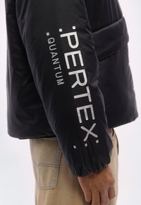 Μανίκι μαύρου μπουφάν τύπου puffer με λευκή επιγραφή "PERTEX QUANTUM", φορεμένο από ένα άτομο με μπεζ παντελόνι, σε α πλό φόντο.