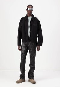 Črna zip-up jakna, siva majica, črne usnjene cargo hlače, bež čevlji. Model nosi refleksna sončna očala; minimalističen moderni dizajn.