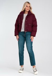 Burgundowa kurtka puchowa z zamkami błyskawicznymi, wysokim kołnierzem i elastycznym dołem. Połączona z jasnoniebieskimi dżinsami i wzorzystymi sneakersami.