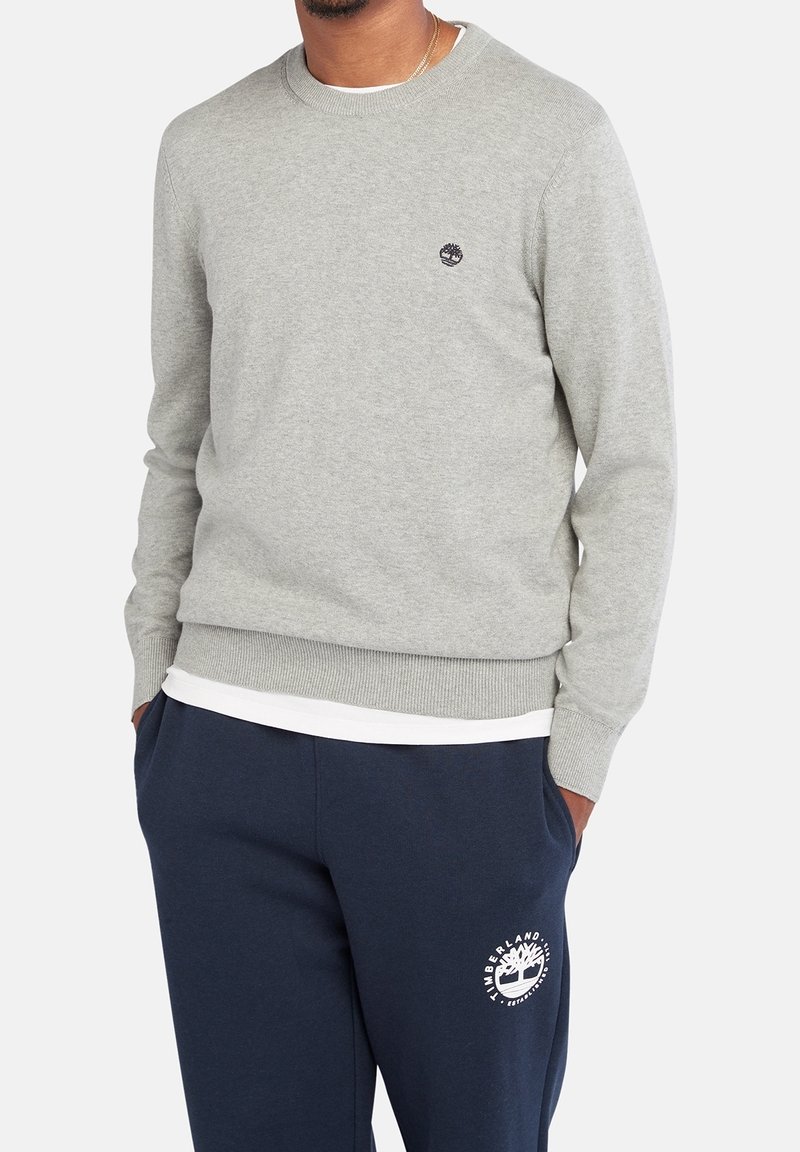 Homme portant un pull gris clair à col rond et un pantalon de survêtement bleu marine avec des logos blancs, debout les mains dans les poches devant un fond blanc.