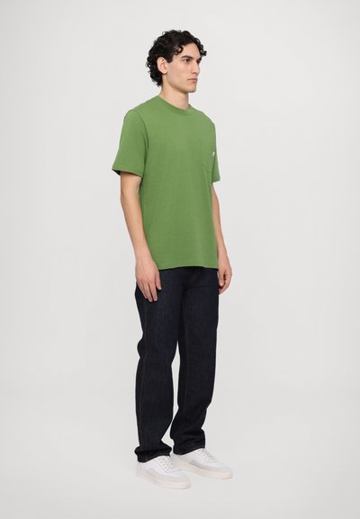 Green cotton t-shirt with a crew neck and short sleeves, featuring a chest pocket. Paired with dark denim jeans and white sneakers.