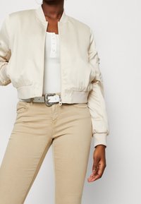 Osoba ubrana w beżową kurtkę bombkę, białą prążkowaną bluzkę z guzikami, beżowe spodnie i biały pasek z ozdobną srebrną klamrą, stojąca z jedną ręką widoczną.