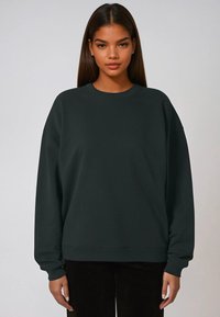 Felpa verde scuro con collo rotondo, spalle scese e polsini a coste. Tessuto morbido con una vestibilità rilassata, abbinata a pantaloni in velluto a coste neri.