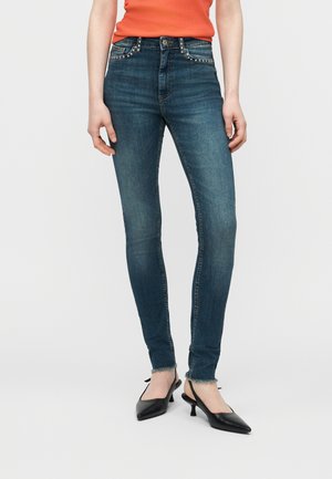 Femme portant un jean slim bleu foncé avec des poches avant cloutées, des ourlets effilochés, des escarpins noirs à bride arrière, et un haut orange, debout sur un fond blanc.